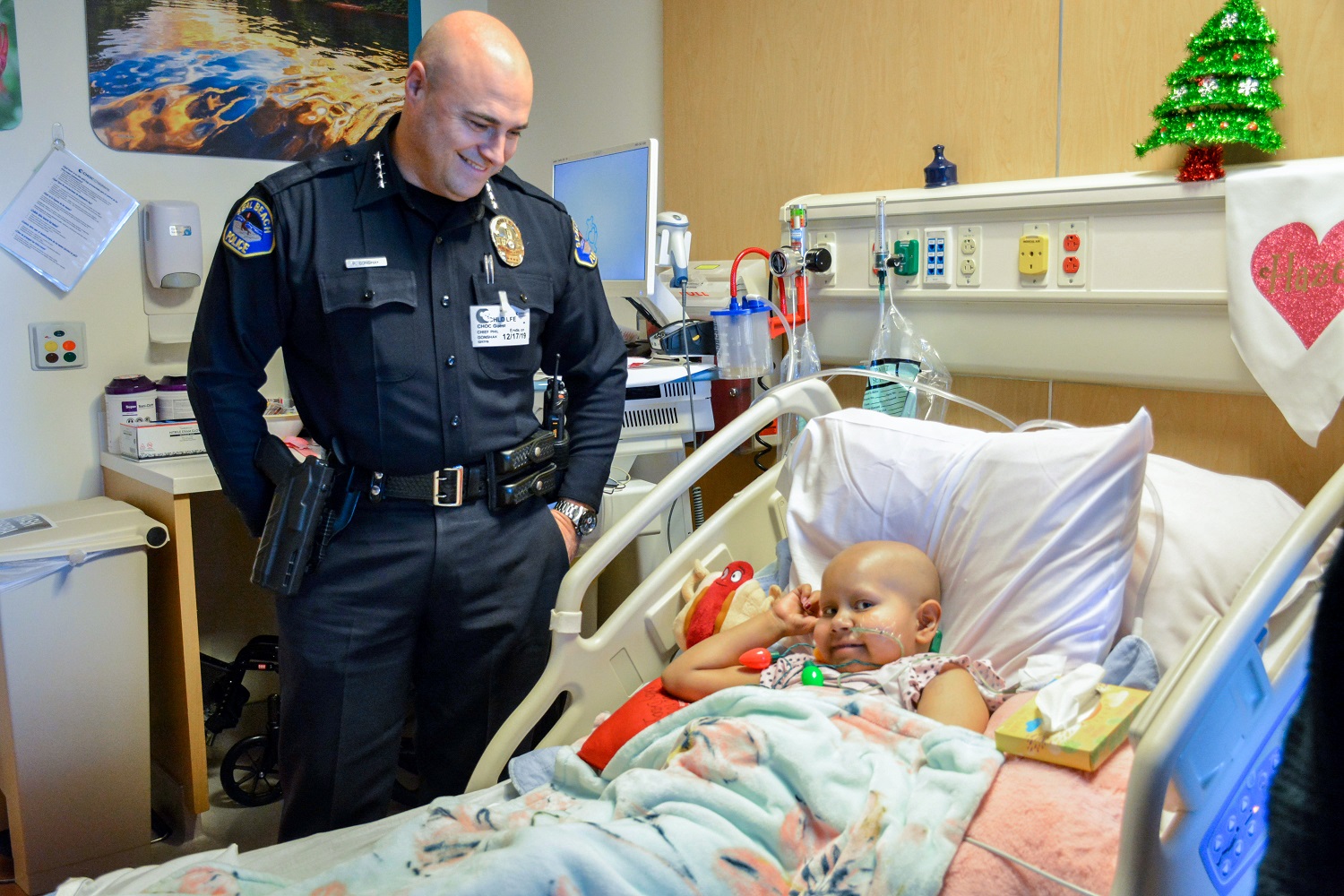 A police officer smiling at a child in a hospital room with holiday decorations; the child is wearing a holiday light necklace
